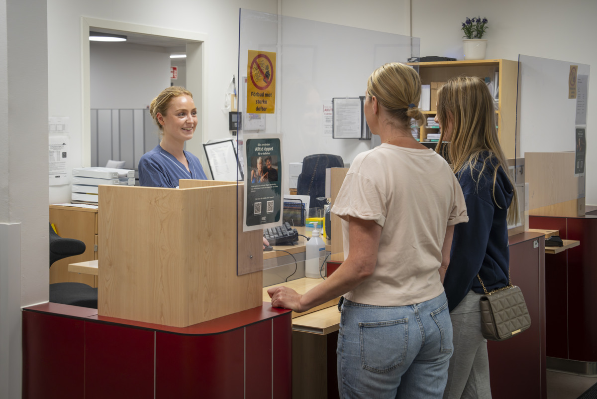 En vuxen kvinna och en ung kvinna som st&aring;r i en reception och pratar med en kvinna i sjukhuskl&auml;der