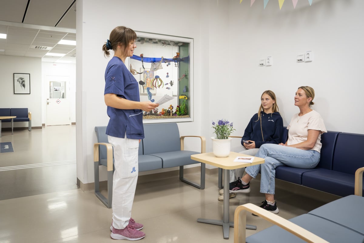 En ung kvinna och en &auml;ldre kvinna sitter i ett v&auml;ntrum och pratar med en kvinna i sjukhuskl&auml;der