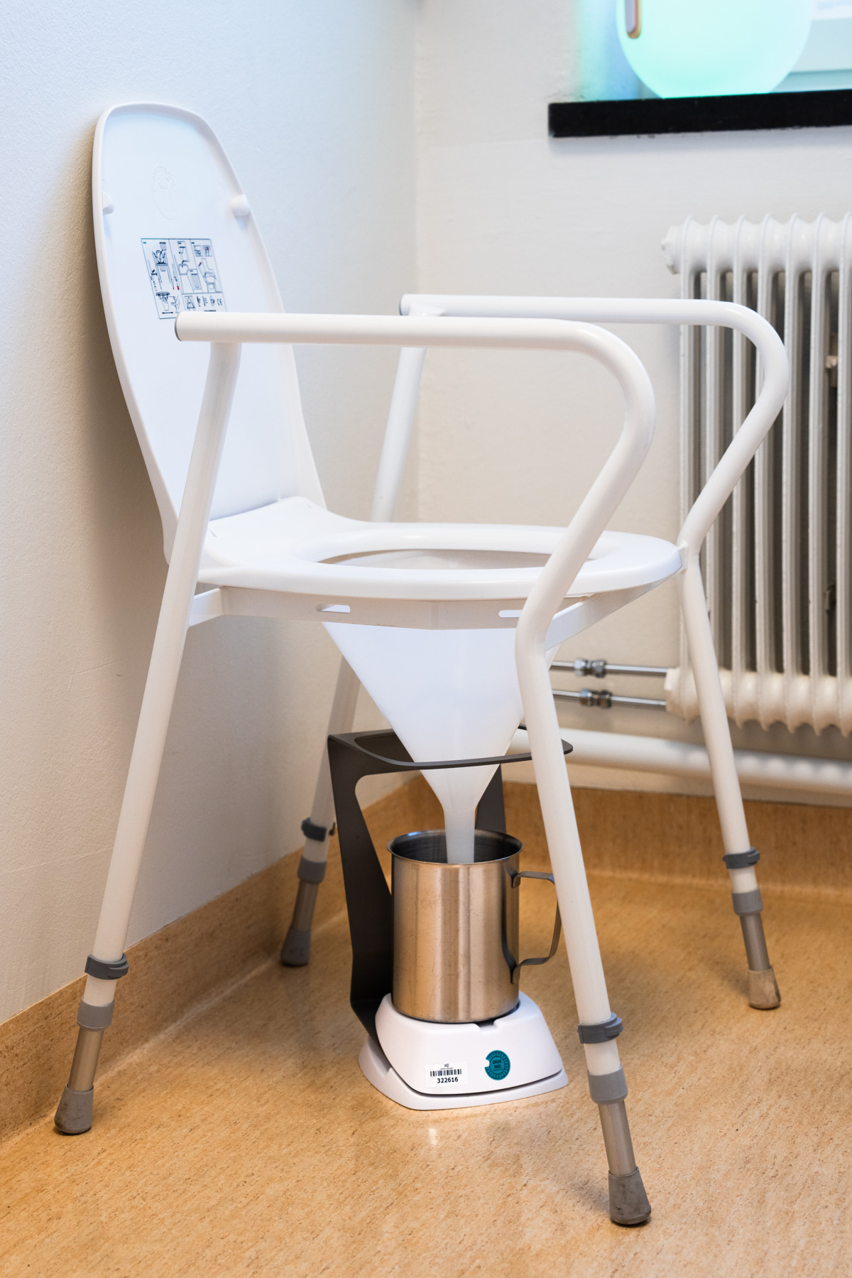 En vit plaststol med en toalettsits och en tratt som leder till en metalbeh&aring;llare 