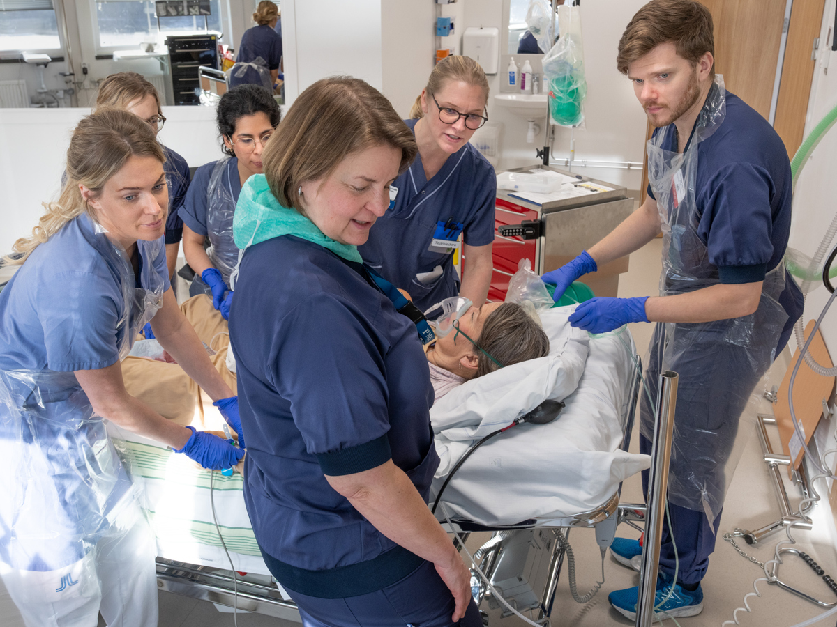 6 personer som utför en övning 5 kvinnor och en man. 1 person som ligger i sjukhussäng och agerar patient. 