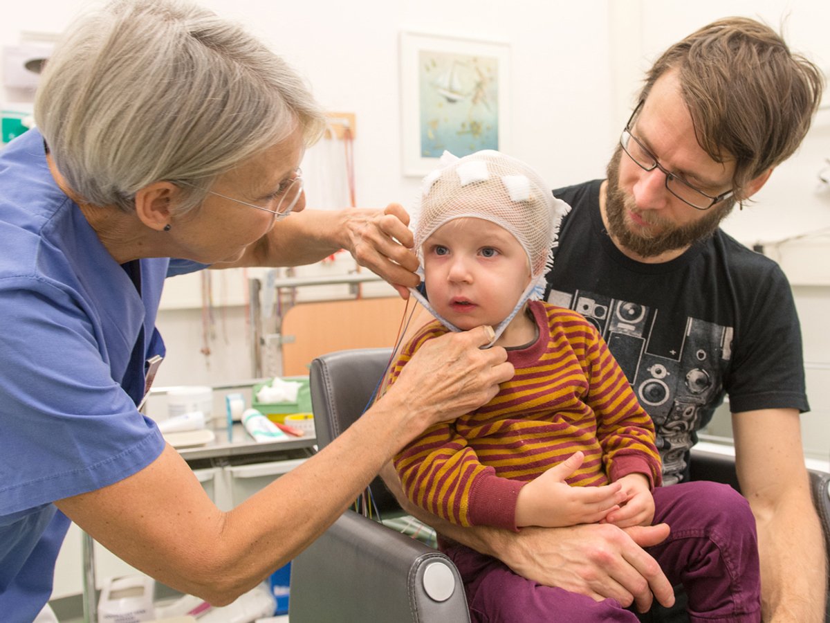 Man med ett barn i sitt knä. Kvinna i sjukhuskläder justerar ett förband som barnet har på huvudet