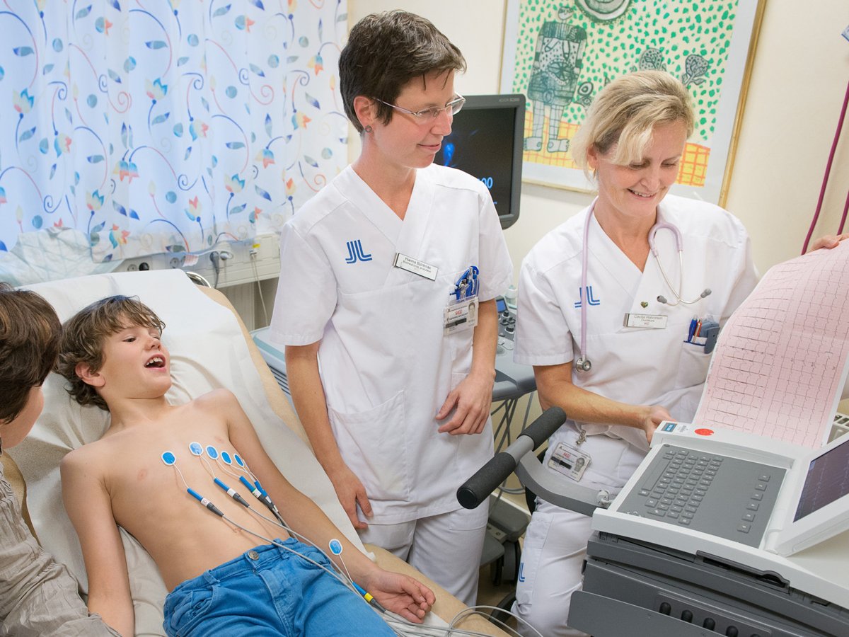 Två kvinnor i sjukhuskläder vid en EKG-maskin, en ung pojke liggandes på en sjukhussäng med elektroder på bröstet och en kvinna bredvid honom