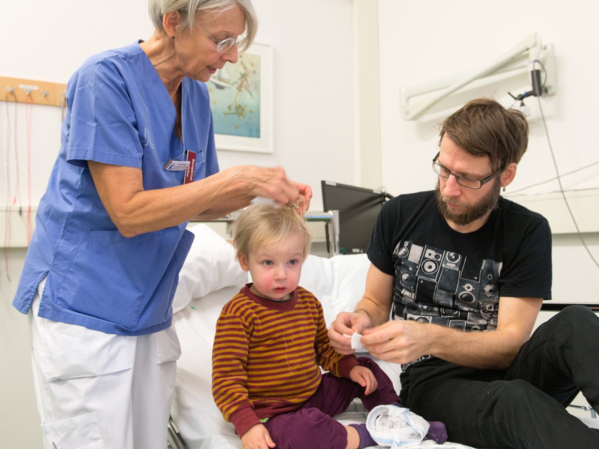Bild på man och barn som ligger i en sjukhussäng och en kvinna i sjukhuskläder som tar bort något från barnets hår.
