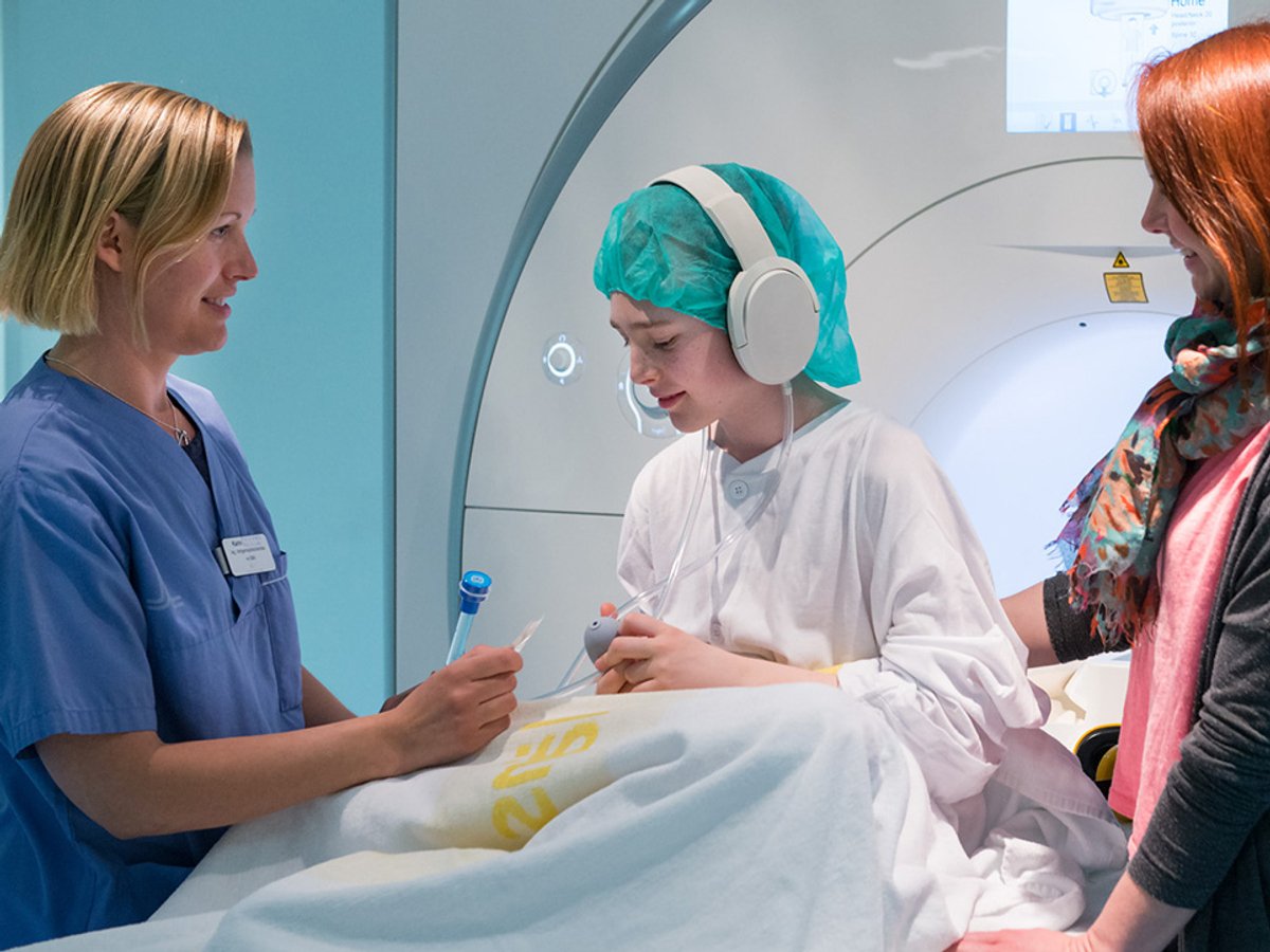En kvinna med sjukhuskläder. En ung pojke som har på sig patientrock, nätmössa och hörlurar på sig. En kvinna med privata kläder. Alla tre står framför en magnetkamera