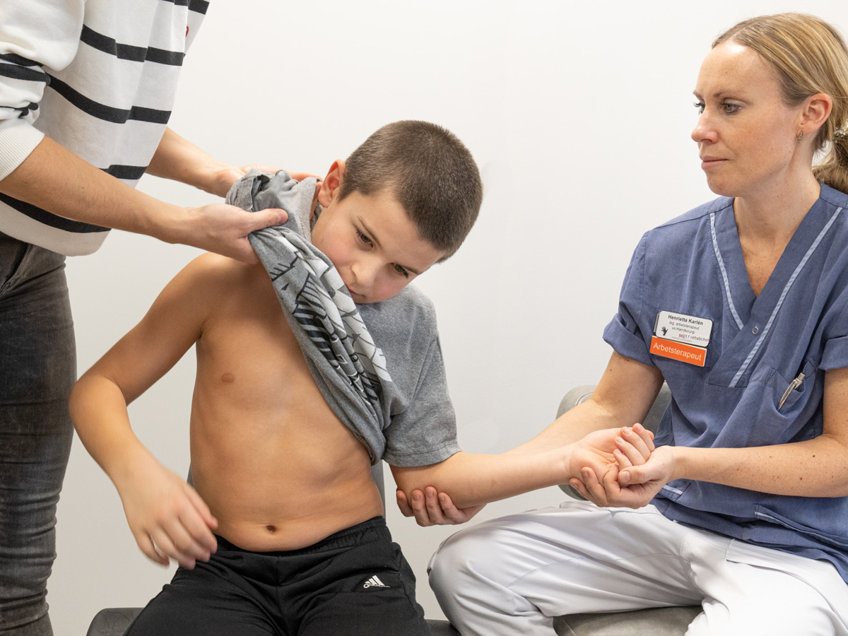 Tre personer. En ung pojke som sitter i mitten. En kvinna i sjukhuskläder till höger om pojken som håller i hans arm. En kvinna till vänster om pojken som hjälper honom att ta på sig en tröja.