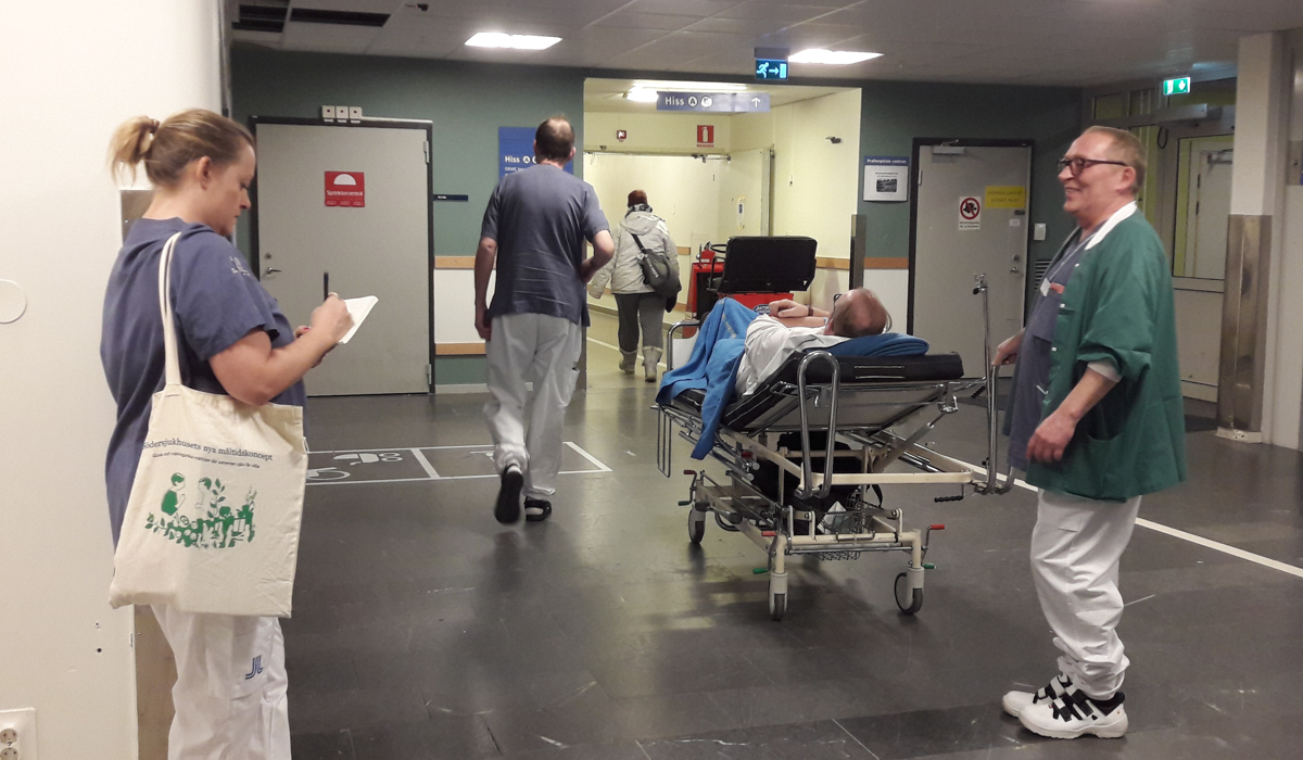 En grupp m&auml;nniskor som st&aring;r runt en patient som ligger i en sjukhuss&auml;ng i en korridor.