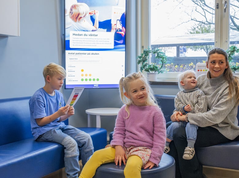 En grupp barn som sitter i ett v&auml;ntrum d&auml;r en sk&auml;rm visar information om hur patienter prioriteras.