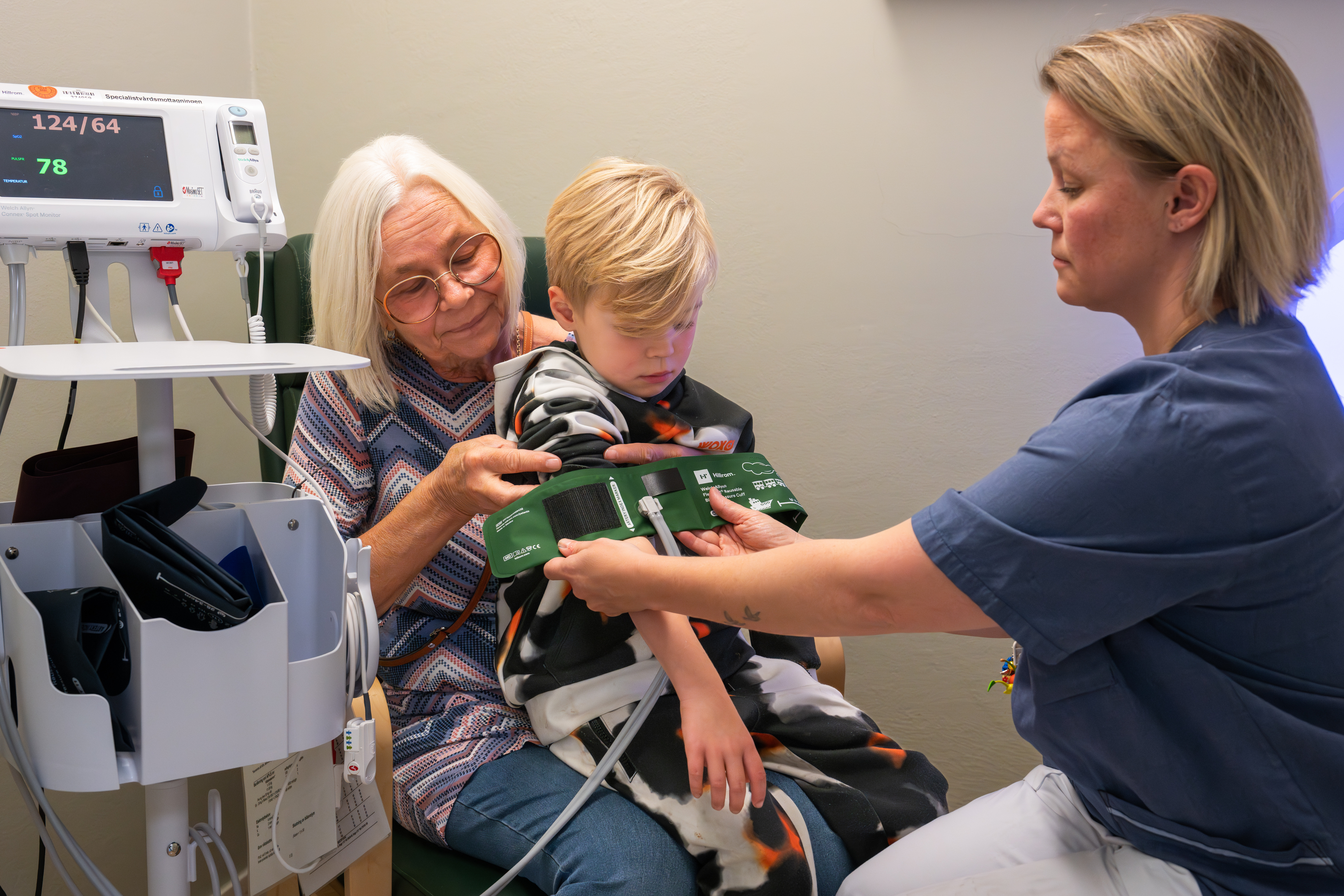 En pojke sitter i en &auml;ldre kvinnas kn&auml; n&auml;r en kvinna i sjukhuskl&auml;der s&auml;tter p&aring; en blodtrycksmanschett p&aring; ett barn