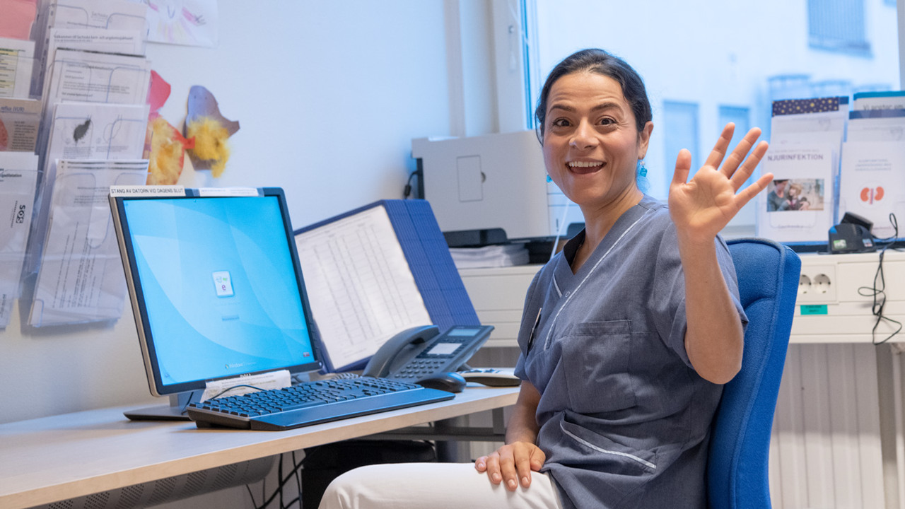 En leende kvinna i sjukhuskl&auml;der sitter  p&aring; en stol och vinkar