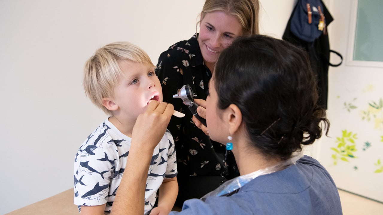 En kvinna i sjukhuskl&auml;der unders&ouml;ker halsen p&aring; ett barn med en tr&auml;pinne och en lampa