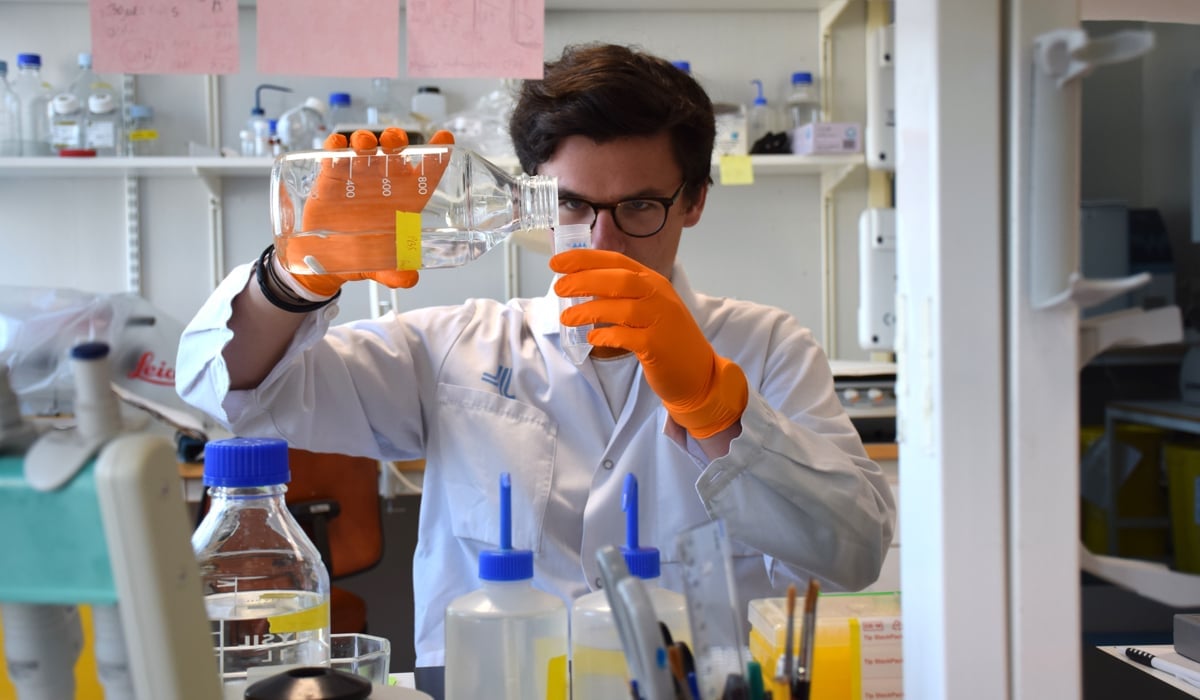 man i vitt labbrock och orange handskar häller vätska ur flaska. 