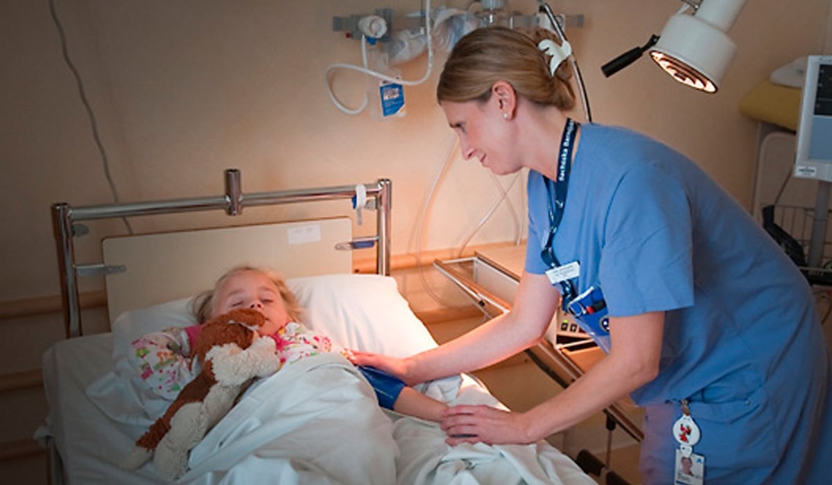 Flicka i sjukhussäng, vårdpersonal lutar sig över sängen och ler