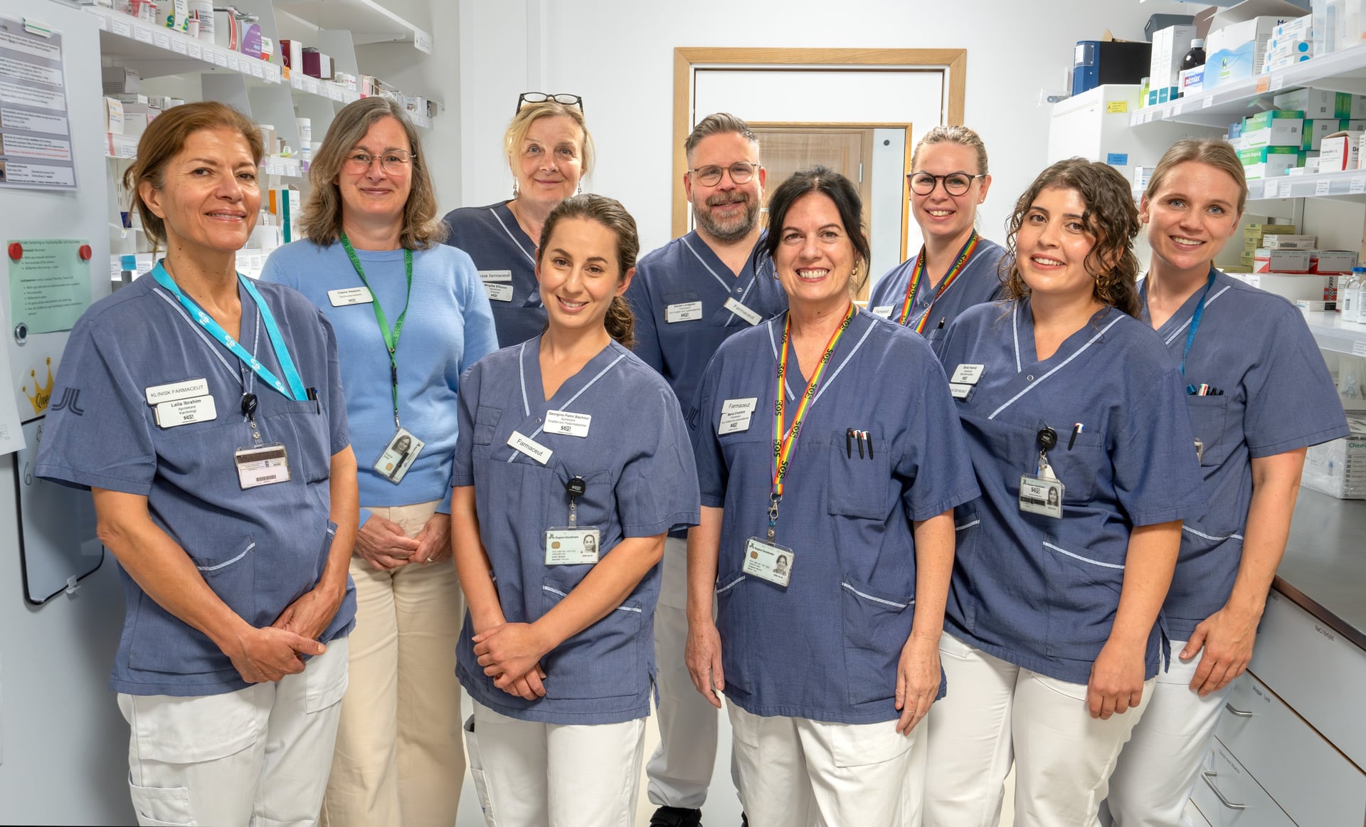gruppbild 8 kvinnor en man sjukhusklädda, läkemedel på hyllorna bakom