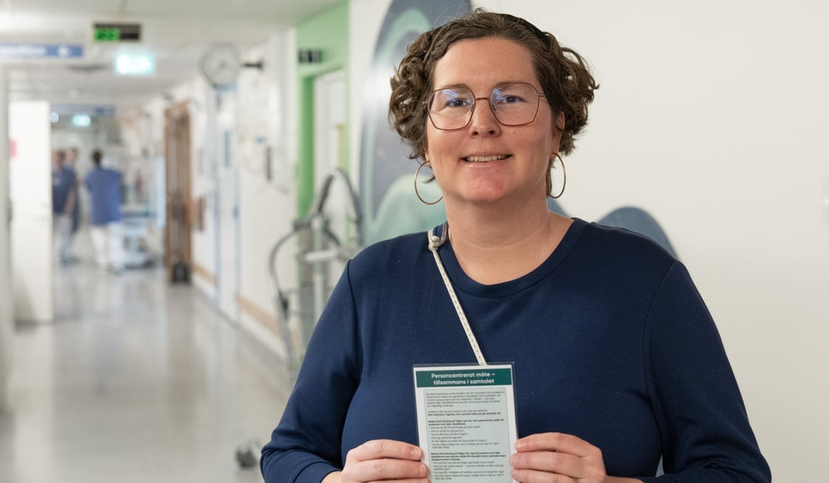 kvinna i blått står i sjukhuskorridor med inplastat kort i händerna.