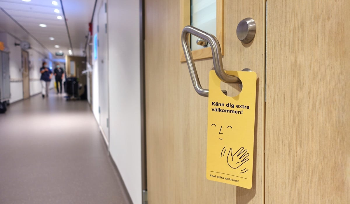 En gul dörrhängare på en dörr i en sjukhuskorridor som visar ett leende ansikte med en hand som vinkar och meningen ”Känn dig extra välkommen”.