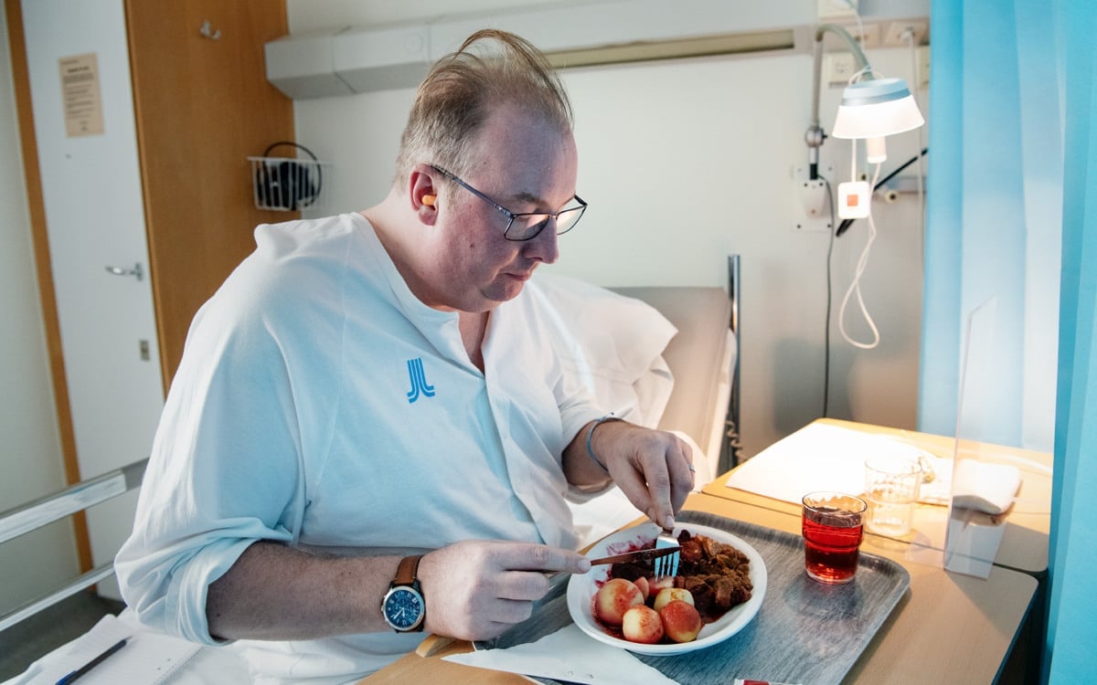 En man i patientkläder sitter i en sjukhussäng och äter mat.