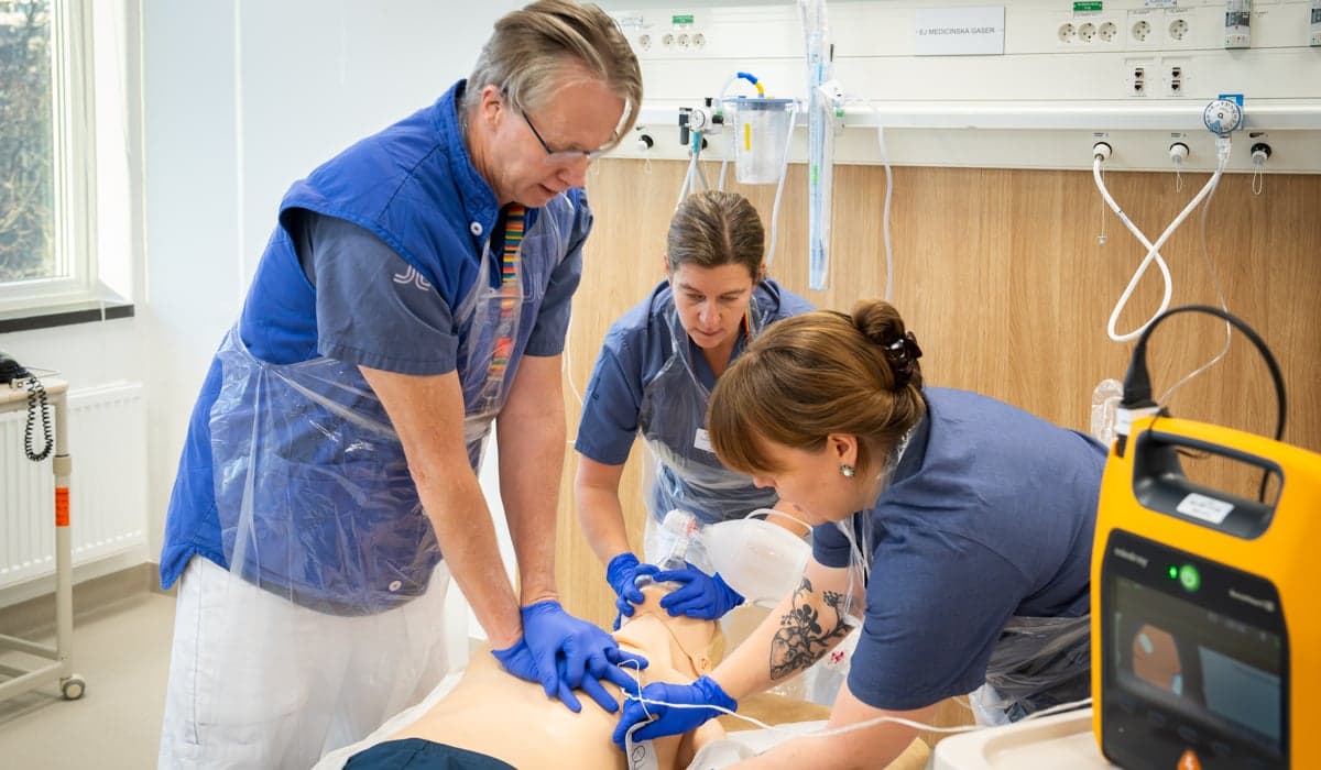 Två kvinnor och en man i sjukhuskläder som övar hjärt- och lungrättning på en docka