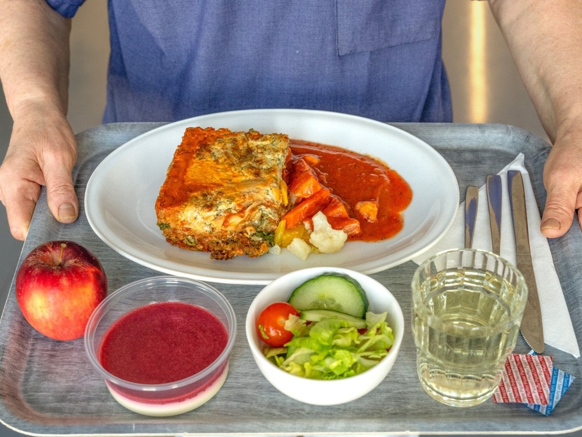 En person med sjukhuskläder som håller i en matbricka med en portion lasagne, en skål sallad, en panacotta, ett äpple och ett glas saft