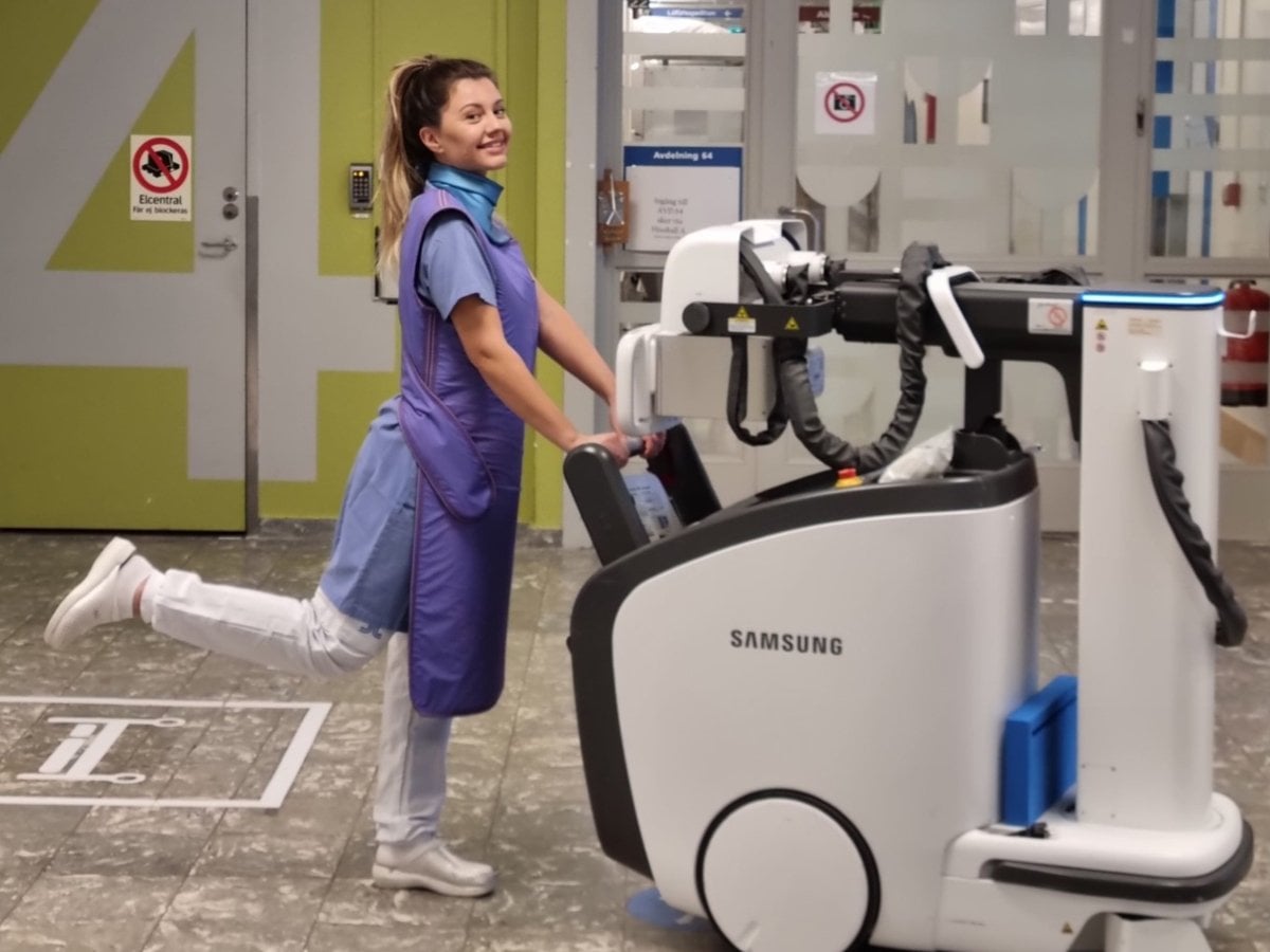 kvinnlig sjuksköterska skjuter röntgenmaskin framför sig i sjukhusmiljö.