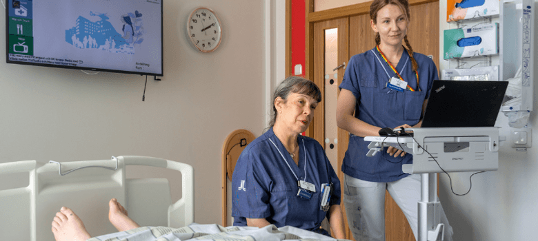 Två kvinnor i sjukhuskläder. En sitter ner och en står upp vid en laptop