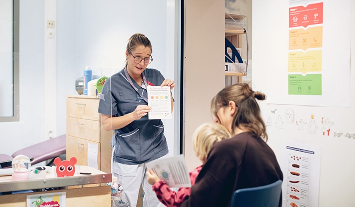 En kvinna i sjukhuskläder som visar ett papper till en kvinna som har ett barn i sitt knä