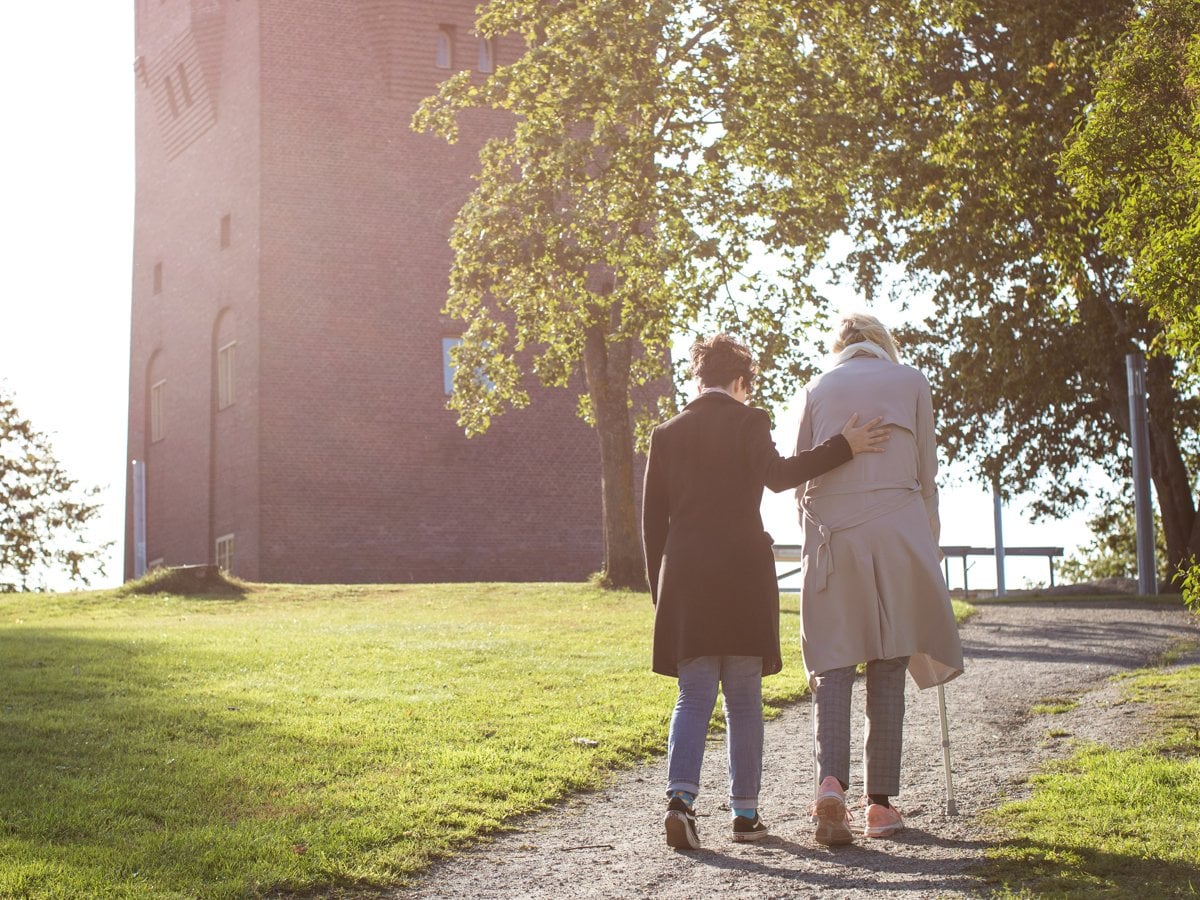 Två personer så går upp för en backe. En med person med kryckor och en annan som håller om personen som går med kryckor.