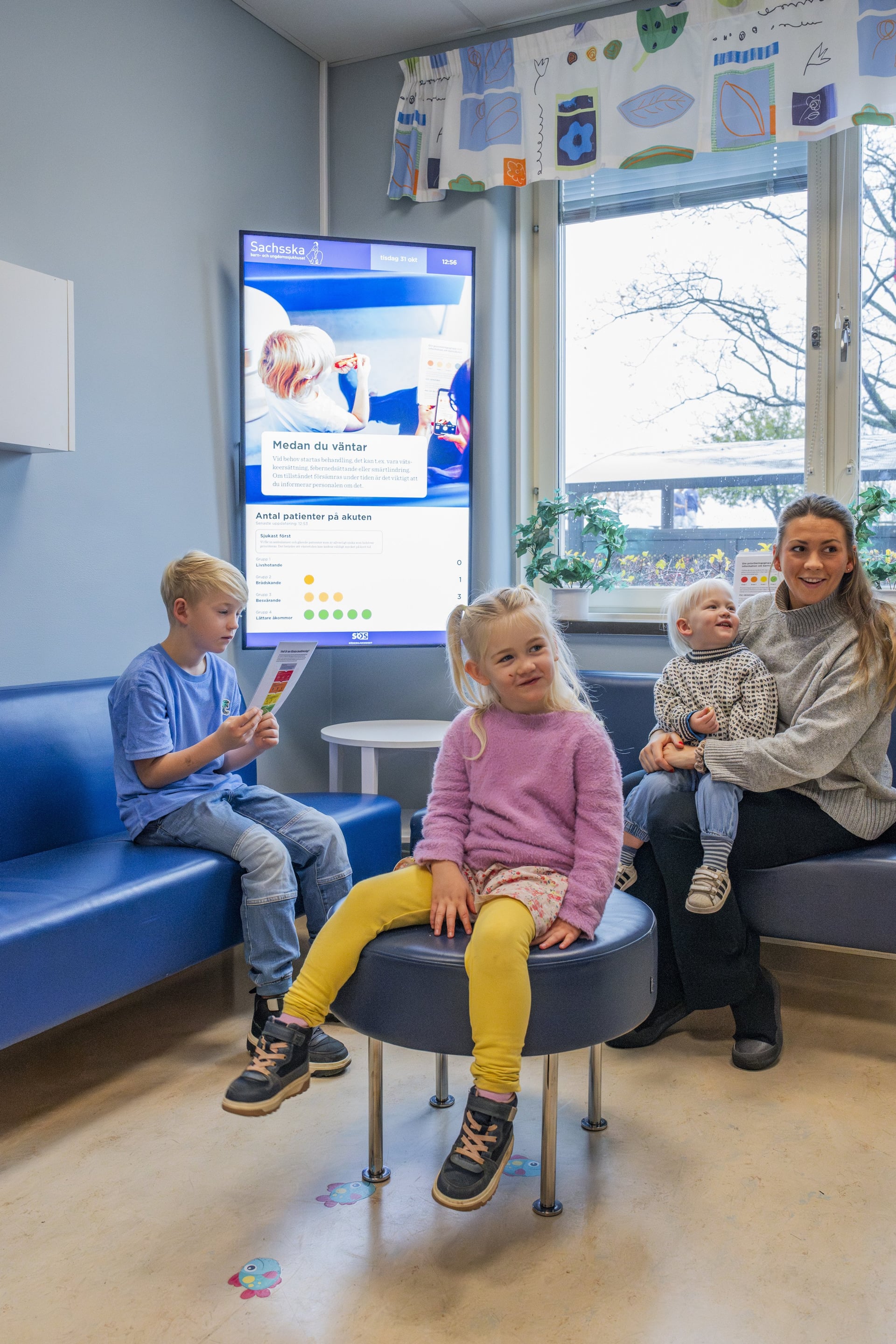 En grupp barn som sitter i ett väntrum där en skärm visar information om hur patienter prioriteras.