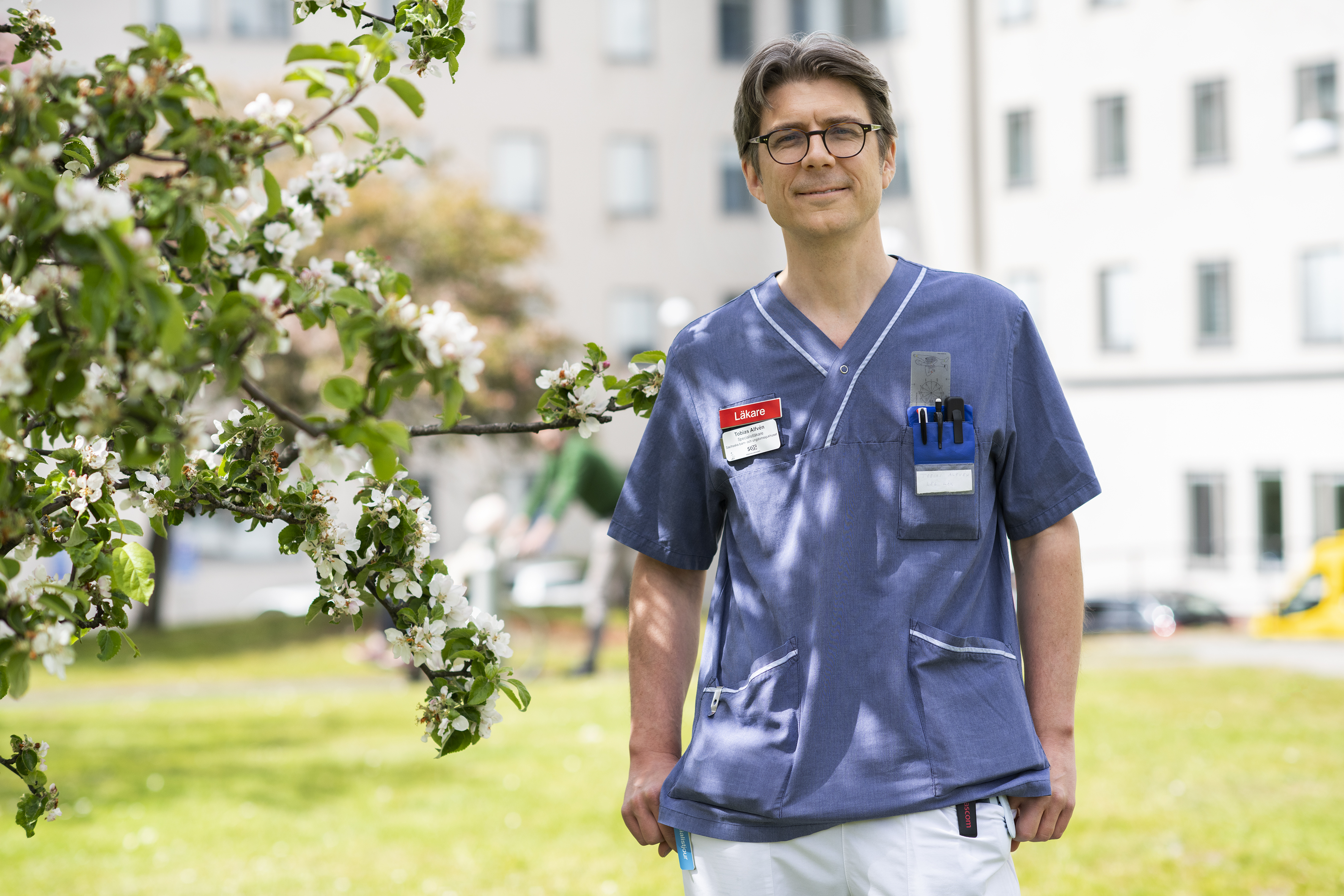 Tobias Alfvén, barnläkare på Sachsska barnsjukhuset, står på en gräsmatta bredvid ett blommande äppelträd.