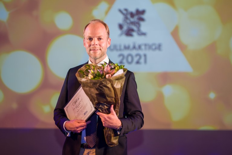 Man stående på scen, håller i blombukett och diplom