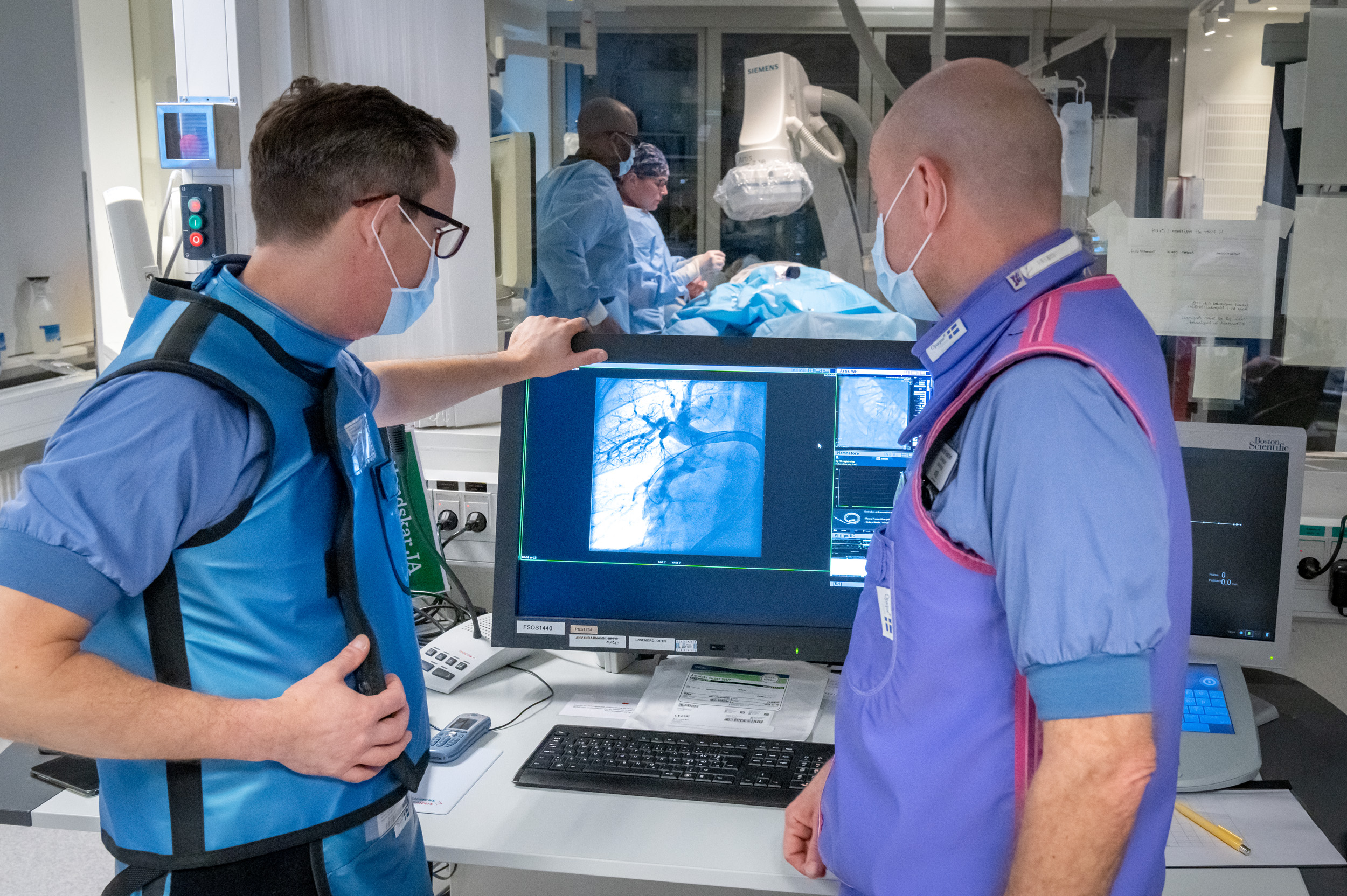 Två personer granskar röntgenbild. I rummet bakom pågår en operation.