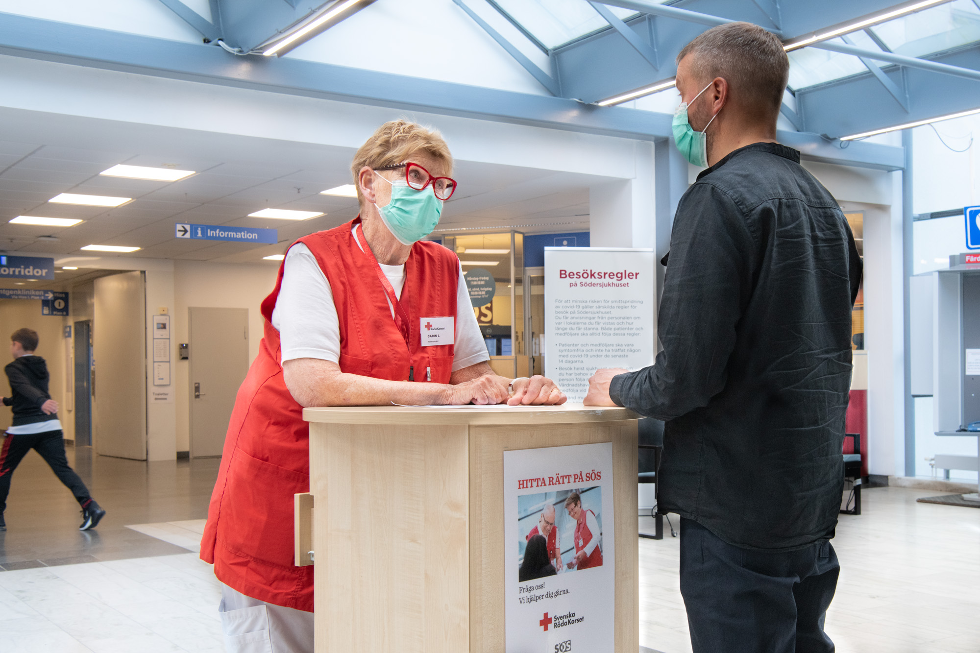 Röda-Korset-värd i röd väst med munskyddstår vid högt bord i Södersjukhusets entréhall och pratar med en besökare i munskydd