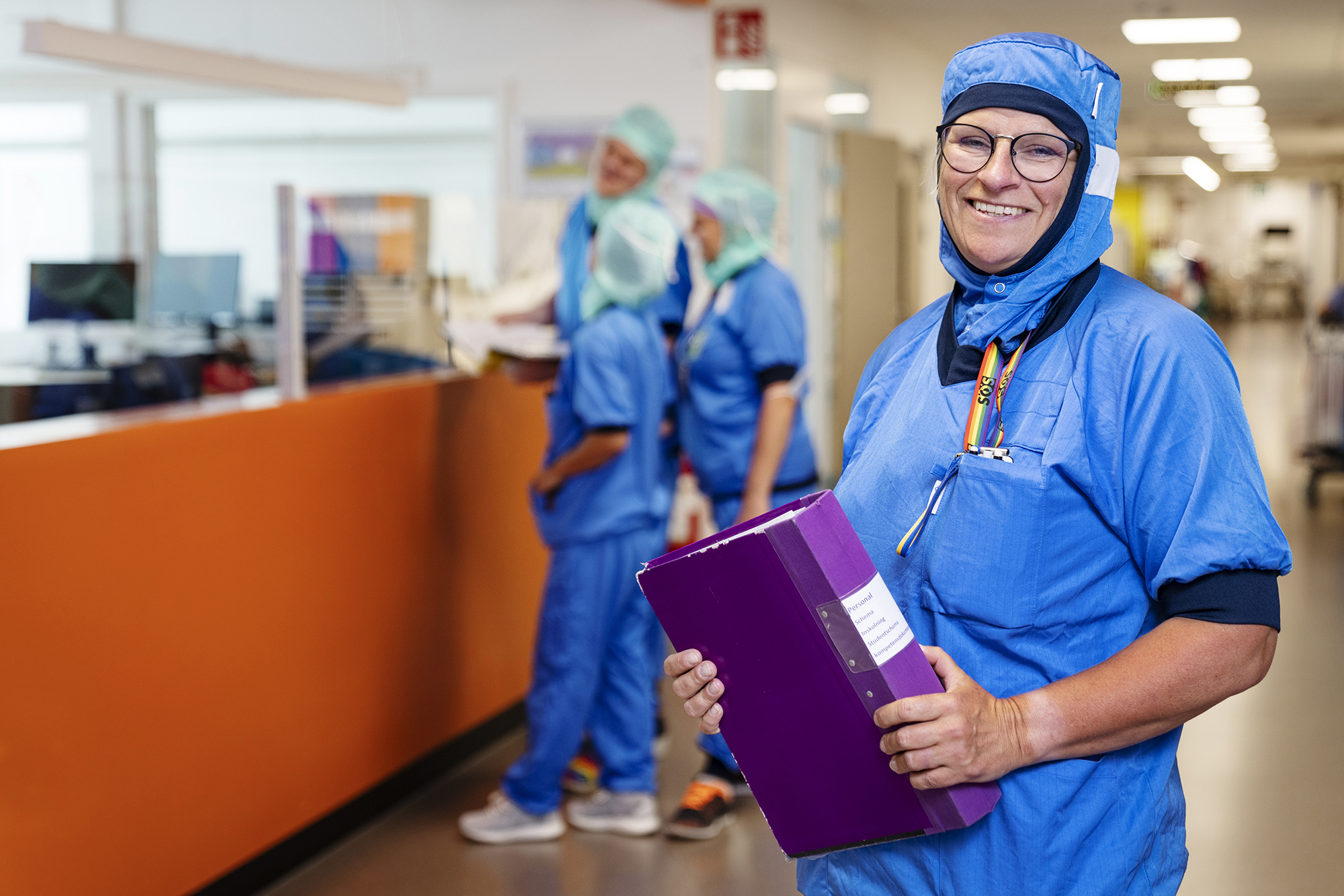 Veronica Dahlgren, vårdenhetschef på ortopedoperation, står i en sjukhuskorridor iklädd blå operationskläder och ler med en lila pärm i händerna.